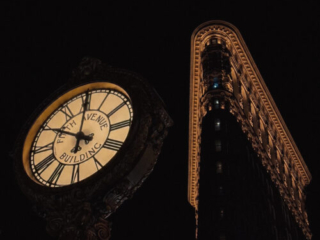 A dramatic night shot looking up at an ornate street clock reading "Fifth Avenue Building" with the illuminated curved corner of the Flat Iron building rising behind it against a dark sky