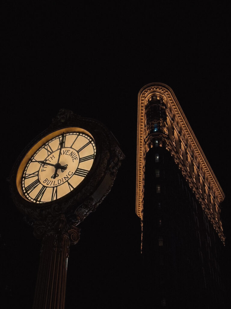 A dramatic night shot looking up at an ornate street clock reading "Fifth Avenue Building" with the illuminated curved corner of the Flat Iron building rising behind it against a dark sky