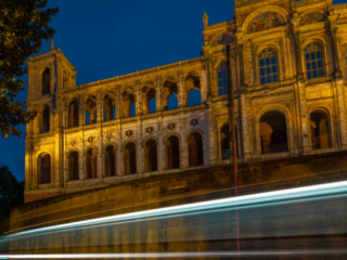 A grand Neo-Renaissance palace or civic building dramatically illuminated in golden light at dusk against a deep blue sky, with long-exposure light trails from passing traffic streaking across the foreground