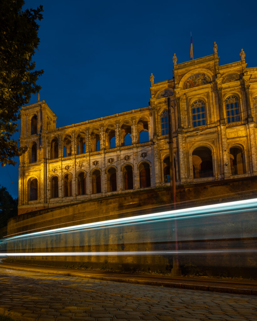 A grand Neo-Renaissance palace or civic building dramatically illuminated in golden light at dusk against a deep blue sky, with long-exposure light trails from passing traffic streaking across the foreground