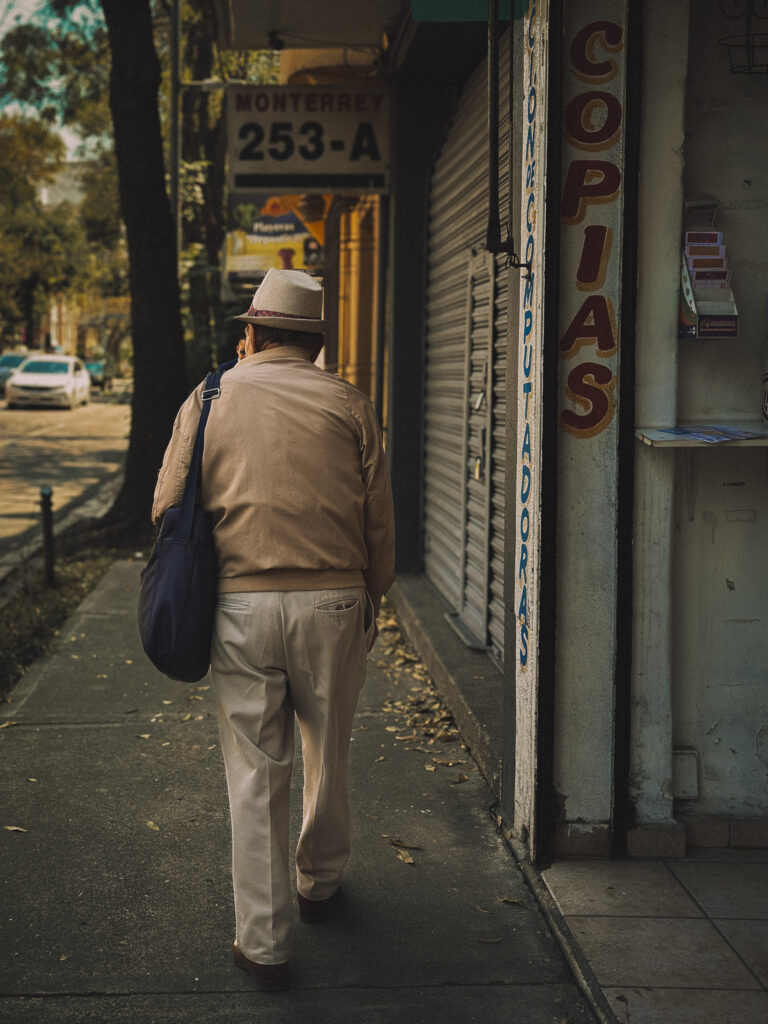 An elderly man seen from behind carrying a blue bag walking down a Mexico City street