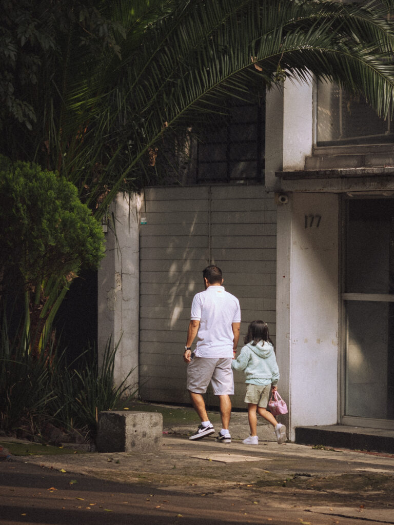 An adult and a young child walking hand-in-hand along a residential pavement, seen from behind, passing a grey building numbered 177 with palm trees above