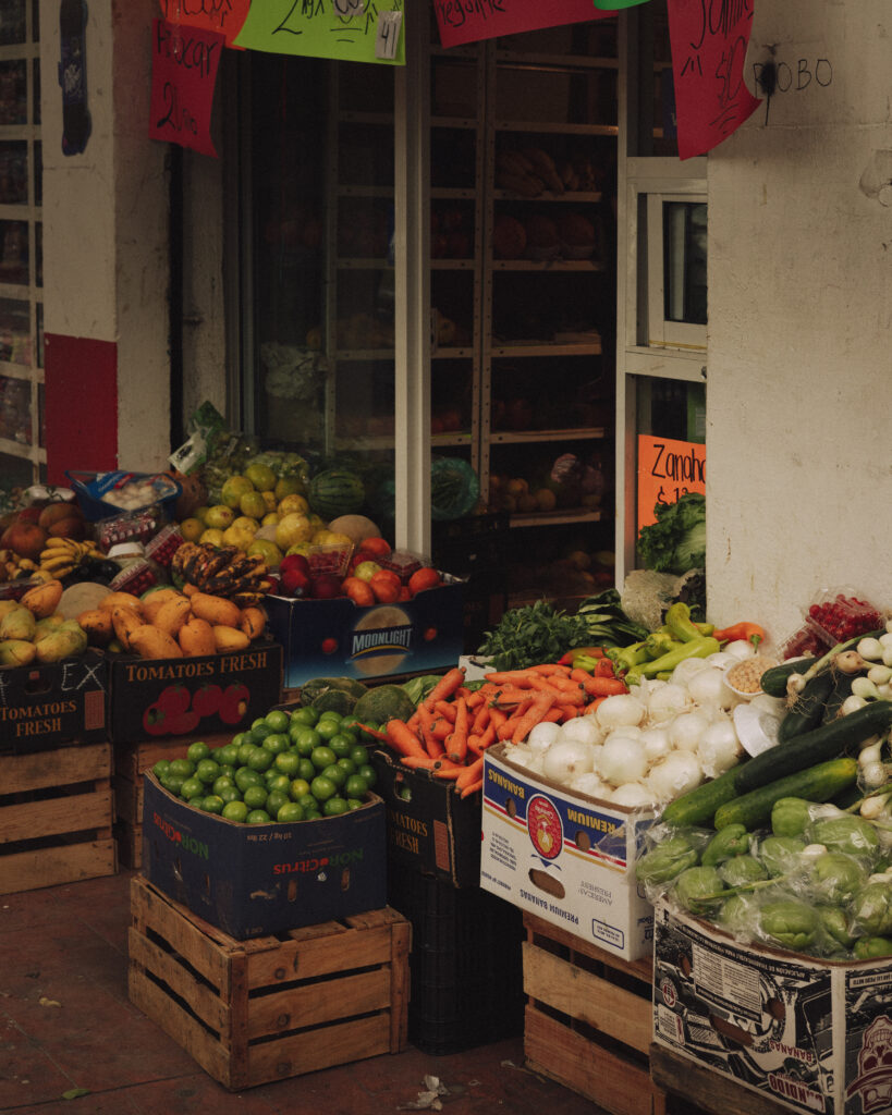A colourful street market stall overflowing with fresh produce including tomatoes, carrots, onions, limes, bananas, and leafy greens displayed in wooden crates and cardboard boxes outside a shop