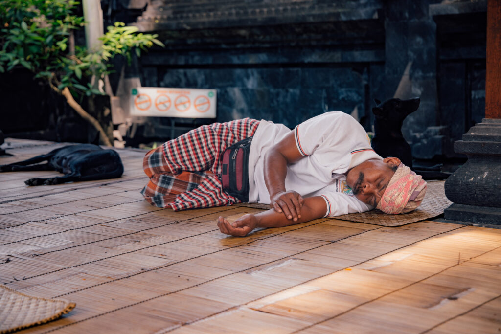 A man in a traditional Balinese headscarf naps on a woven mat at a temple, a dog resting quietly beside him